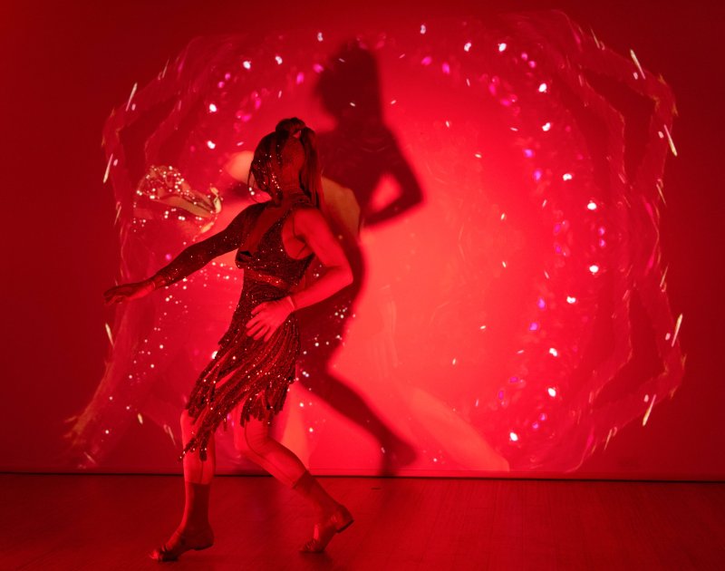 Performer in a sparkly dress and tights dances against a red wall with a shimmering, projected light pattern and a large shadow.