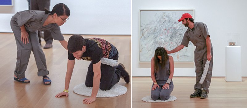 Two people in a gallery, one on a rug on all fours, the other standing; a man with a red hat touches a woman kneeling on a rug.