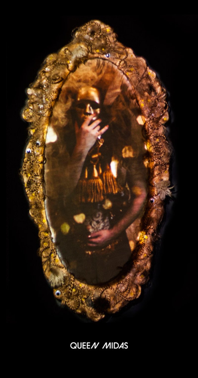 Ornate gold mirror reflecting a figure with a gold mask, long hair, and gold tassels, on a dark background.