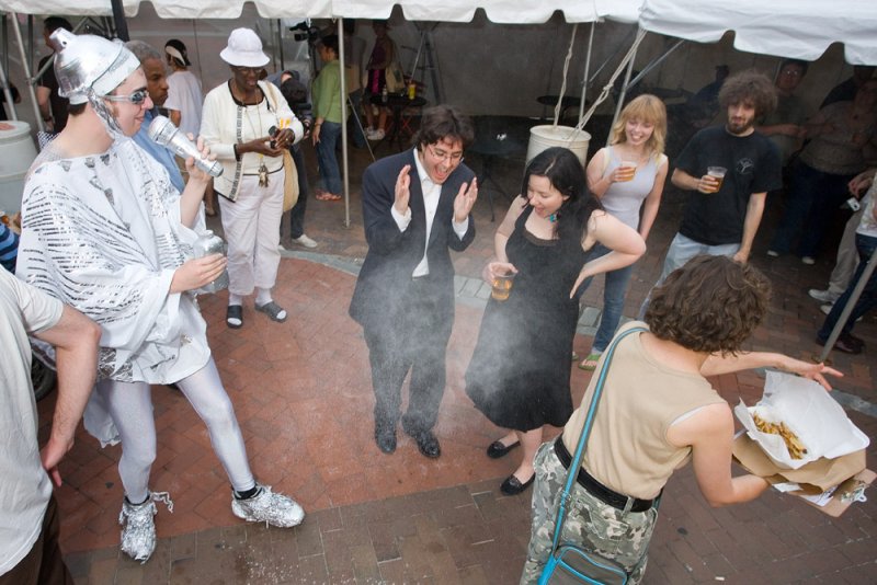Performance art with a man in a silver helmet and robe spraying mist, surrounded by laughing onlookers.