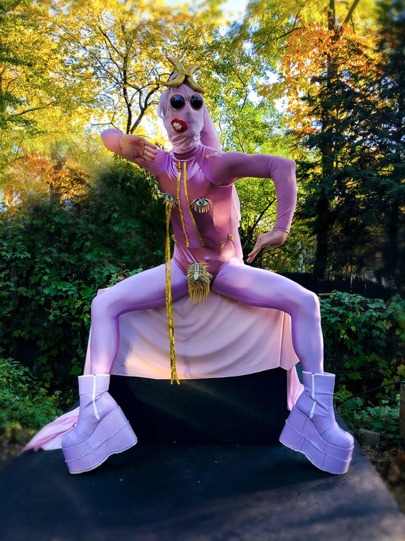 A person in a full-body pink suit, mask, and platform boots, posing outdoors with trees in the background.