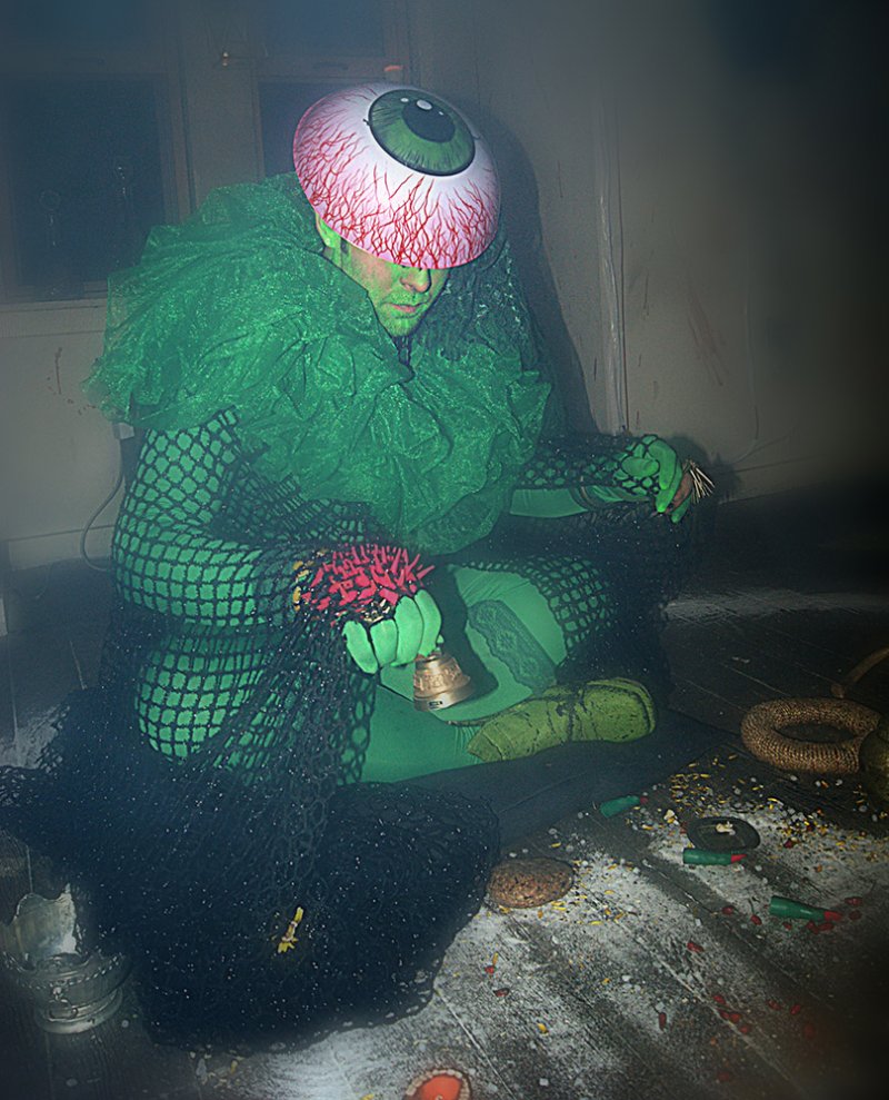 A person in a green costume with a giant eyeball mask, sitting on a dark floor covered in white powder and small objects.