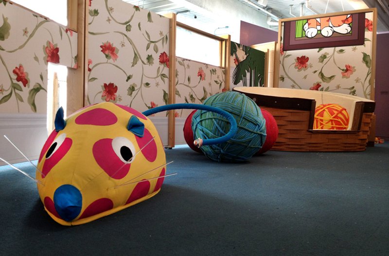 Installation view featuring a large, yellow, polka-dotted mouse-like form connected by a blue tube to a large ball of yarn. Floral wallpapered partitions and a cat-themed structure are in the background.