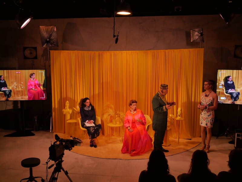 Performance art with four costumed figures on a stage with a bright yellow curtain, two screens on either side.