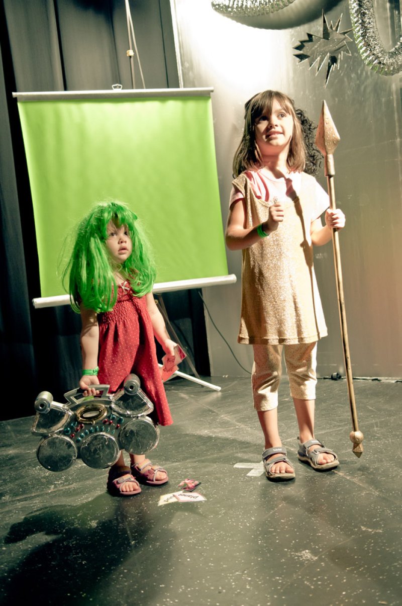 Two young girls in costumes on a stage. One in a green wig and red dress holds a toy drum set; the other in a gold dress hold