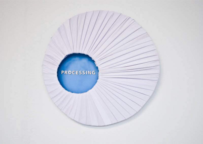 White pleated fabric circle with a blue center displaying "PROCESSING" in white text, mounted on a white wall.