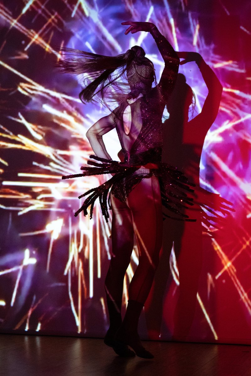 Dancer in a sparkling costume with a fringed skirt, silhouetted against a vibrant red and purple background with bright, exploding light patterns.
