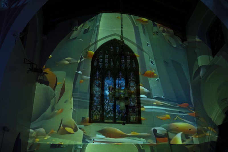 Digital projection of fish swimming around a gothic stained-glass window, casting a green glow on a dark church wall.