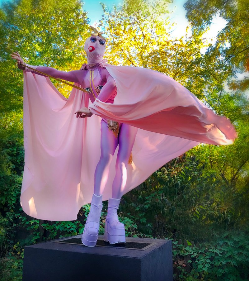 Performance artist in a pink bodysuit, cape, and platform boots, with a masked face, standing outdoors.
