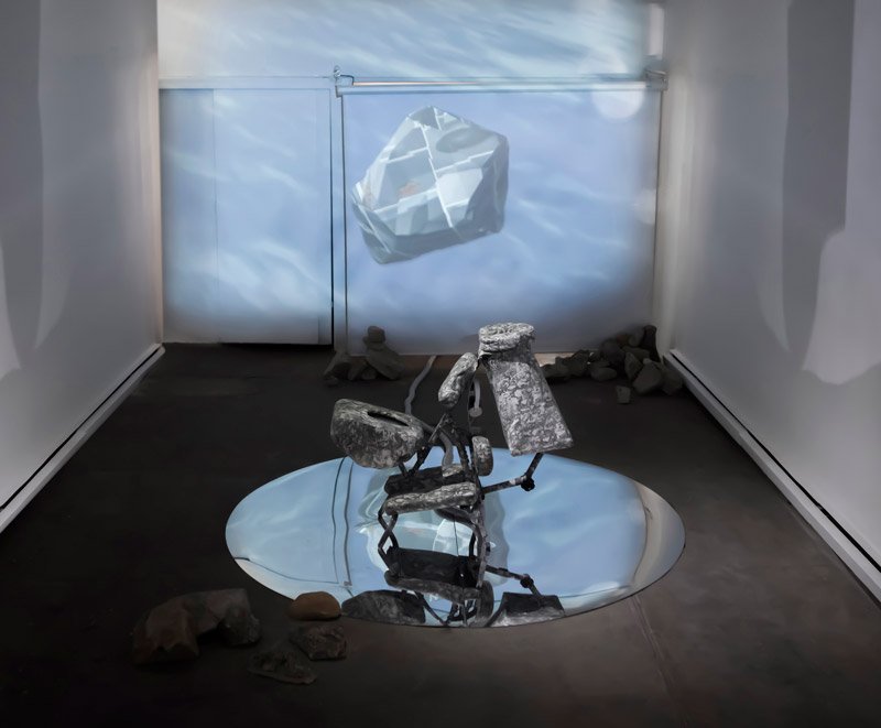 Installation with a mirrored oval on a dark floor reflecting a grey sculpture, and a projected image of a floating rock on the back wall.