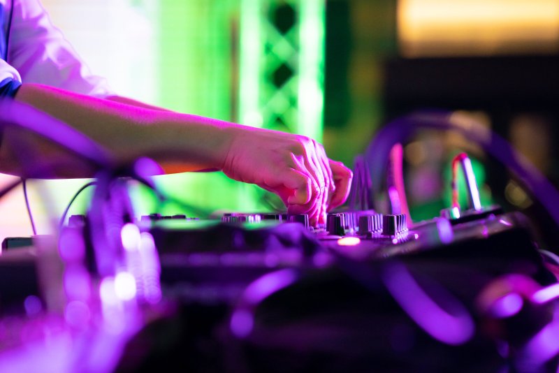 Close-up of a DJ's hands on a mixer, illuminated by vibrant pink and purple lights, with blurred green lights in the backgrou