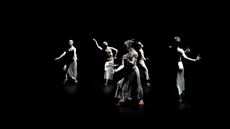 Five dancers in white skirts and painted torsos pose in a dark space, illuminated by spotlights.