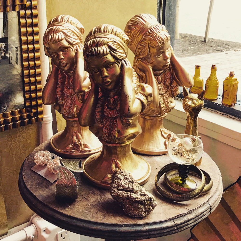 Three golden busts of women with hands to their ears on a wooden table, with a golden cat figurine and bottles.