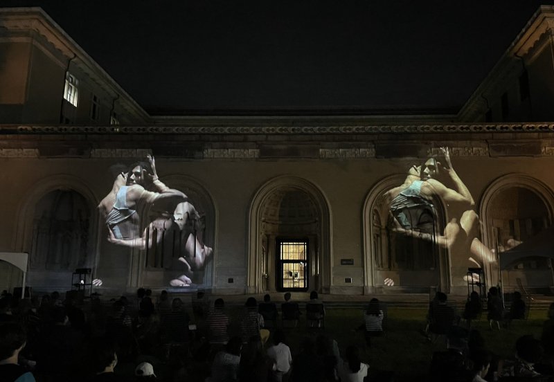 Night projection of two large, light-toned figures on a dark stone building, with an audience seated below.