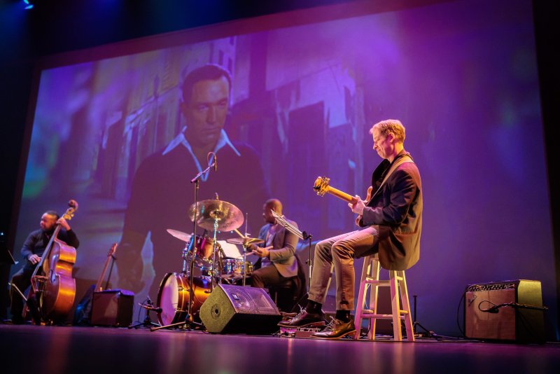 Scott N Andrew's "Suite Life" (2022-2023) shows a live band on stage with a large projection of a man's face behind them.