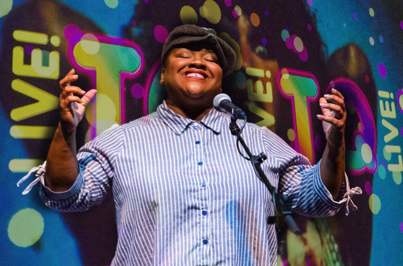 Smiling person in a striped shirt and cap stands at a microphone, with hands raised. Behind them, a colorful backdrop features the word "LIVE!" in large, outlined letters.