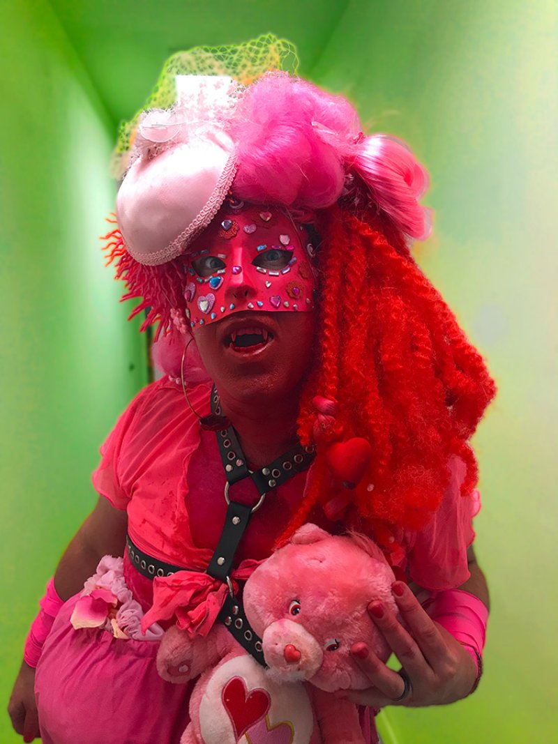 Person in vibrant pink costume with red face paint, fangs, elaborate wig, and a pink teddy bear.