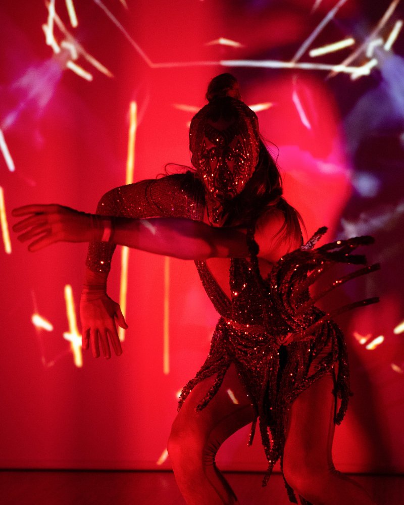 Dancer in a sparkling red costume and mask, arms outstretched, against a vibrant red background with abstract light projections.