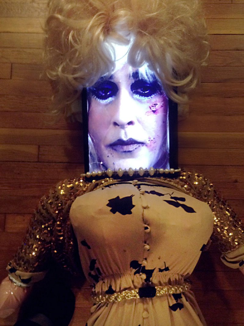 Drag queen's face projected onto a screen, topped with a blonde wig, on a mannequin in a sequined dress.