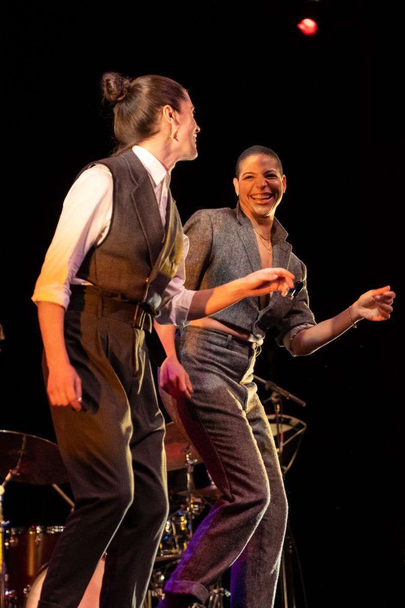 Two women in suits dance on a stage with dark background, one smiling broadly, the other looking left.
