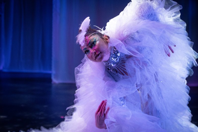 Dancer in a ruffled white costume with elaborate face glitter and a feathered headpiece, illuminated by blue and purple stage lights.
