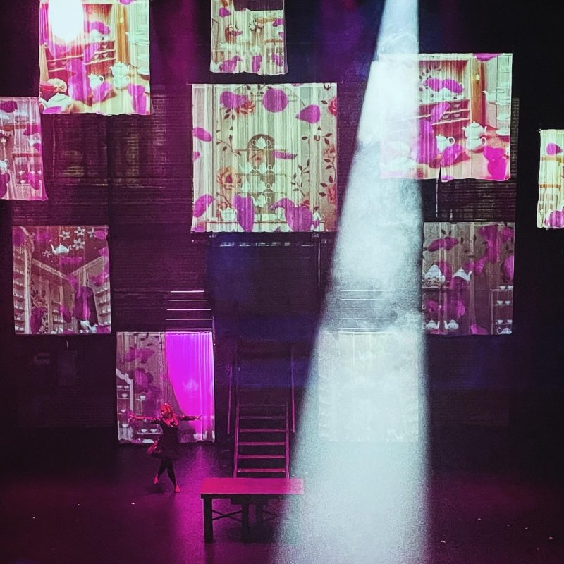 Stage view with a performer, table, and stairs under a spotlight. Backdrops show pink floral patterns and teacups. The scene is lit with purple and white light.