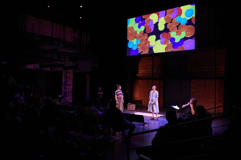 Theater scene with two figures in colorful dresses on stage, facing the audience. A screen above displays an abstract pattern of colorful spheres. Brick walls and audience in the dark background.