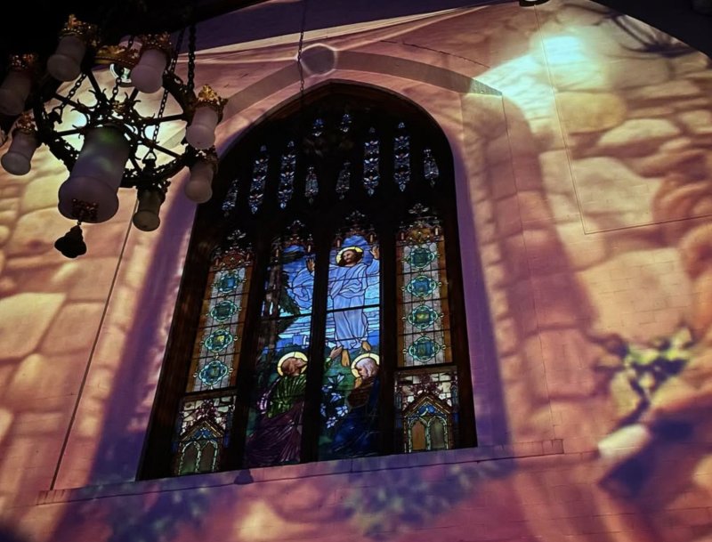 Stained glass window depicting Jesus and two figures, illuminated by a chandelier and projected light creating a textured, pinkish glow on the wall.