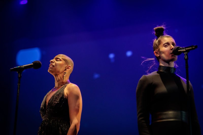 Two singers, one bald with a dark patterned dress, the other with hair in a bun and a black outfit, singing into microphones on a blue stage.