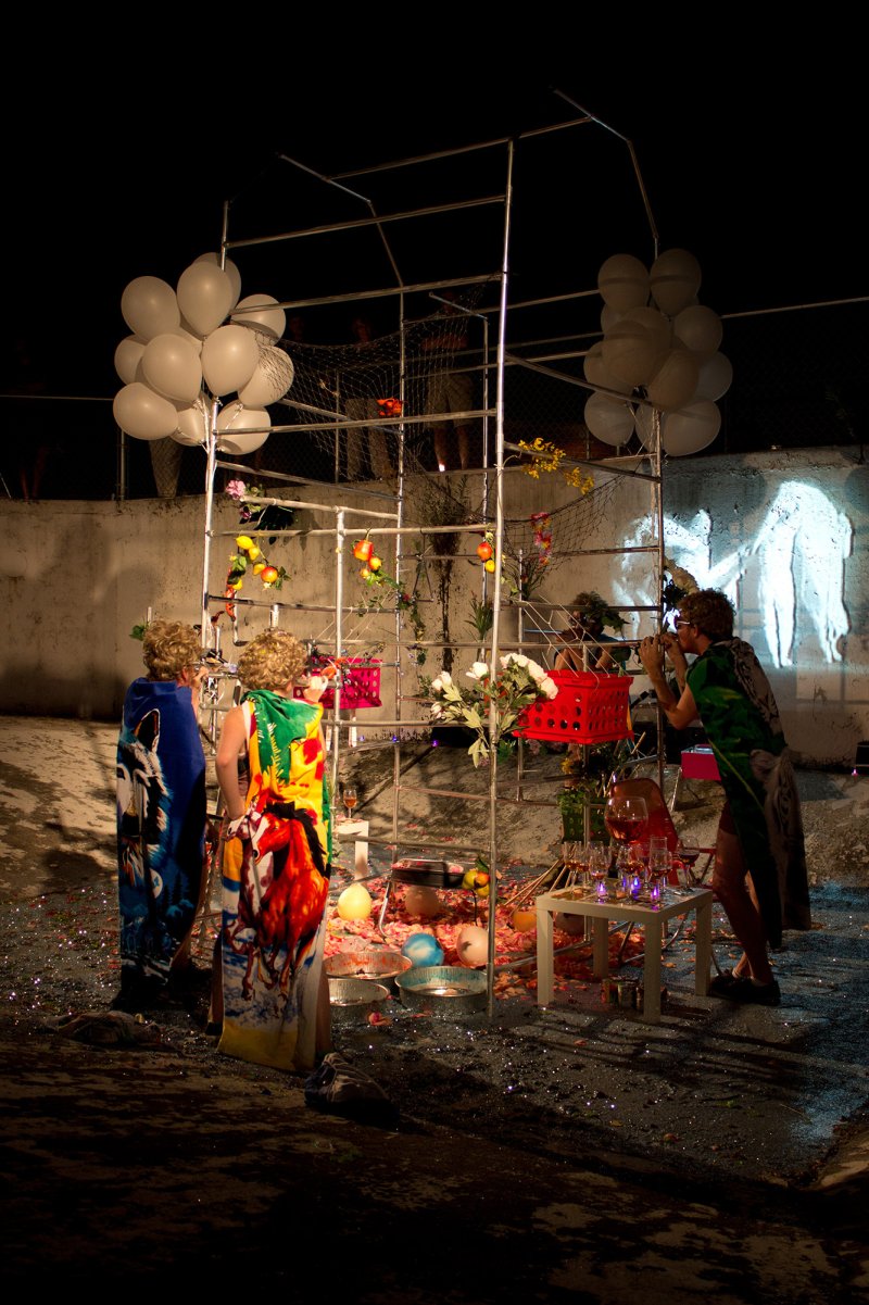 Scott N Andrew's "Gutai Beach Party" (1772359854) shows three figures in colorful robes near a metal structure adorned with balloons, flowers, and objects, with a projected image o