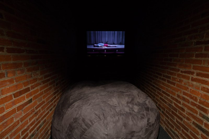 Dark, brick-lined tunnel with a large gray beanbag chair. A screen at the end shows a person lying on a stage.