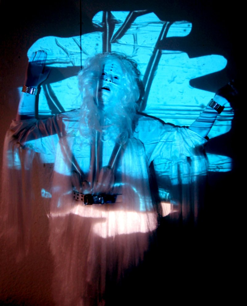 Performance art with a person in white, long-sleeved clothing and a white wig, illuminated by blue light.
