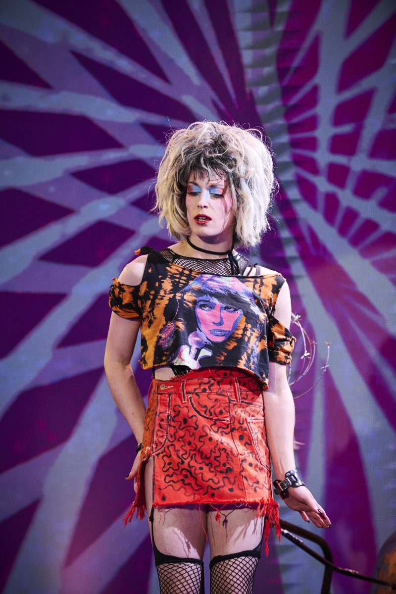 Drag performer in blonde wig, ripped graphic tee, orange skirt, fishnets, and bold makeup against a purple geometric background.