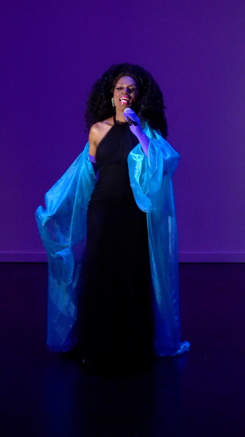 Drag queen in a black gown and flowing blue cape, singing into a purple microphone under purple stage lights.