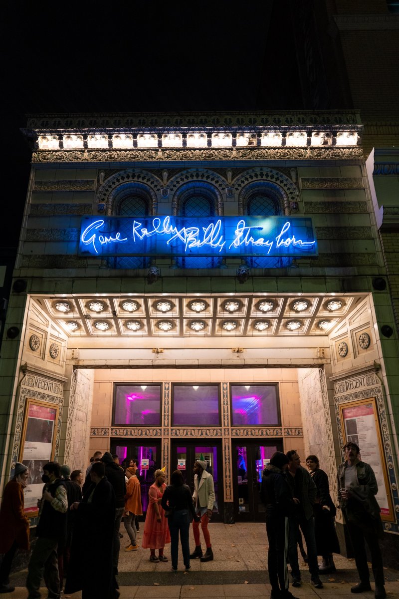 Night shot of ornate building entrance with blue neon sign, crowd of people, and purple interior lighting.