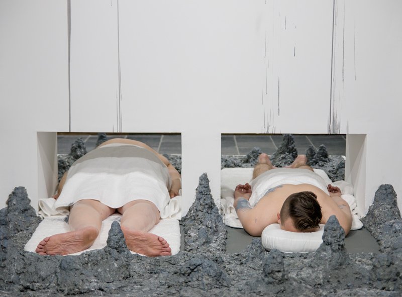 Two people lie face down in grey, rocky alcoves. One is pale with bare legs, the other has tattoos and is covered by a white sheet.