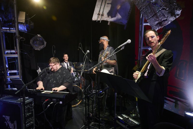 Four musicians on a dark stage with bright lights. A man on keys sings, another plays guitar, a drummer smiles, and a bassist plays.