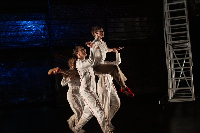 Three dancers in white jumpsuits and red-striped shoes, one held aloft, against a dark stage with blue lighting and a ladder.