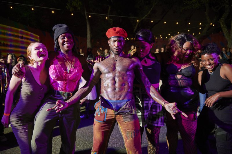 Five people pose at night, one shirtless man with a red beret and unzipped pants at the center.