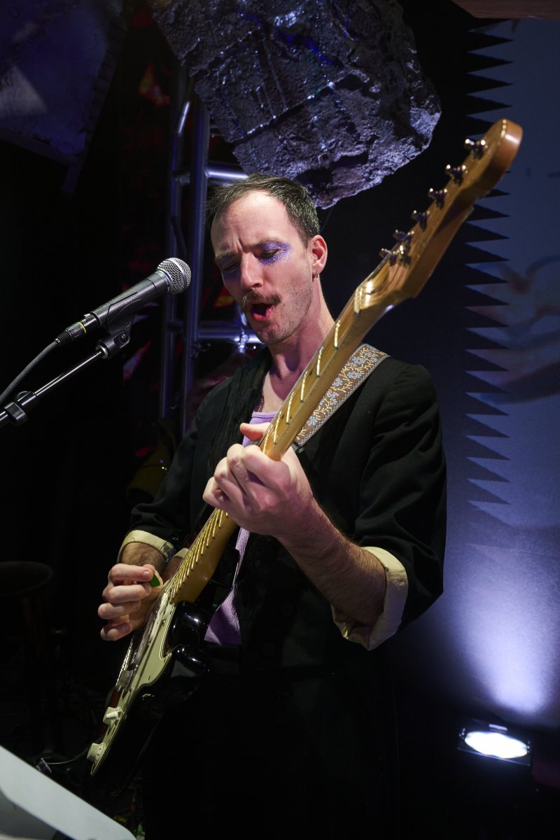 Man with purple glitter eyeshadow and open mouth sings into a microphone while playing an electric guitar with a patterned strap.