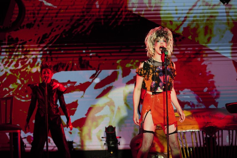 A person in drag with a blonde wig, orange skirt, and fishnets sings into a microphone on stage, with a bandmate and red-patterned backdrop.