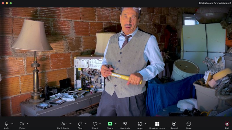 Man in vest and tie, with safety glasses on head, holds a yellow mallet in a cluttered room with brick walls.
