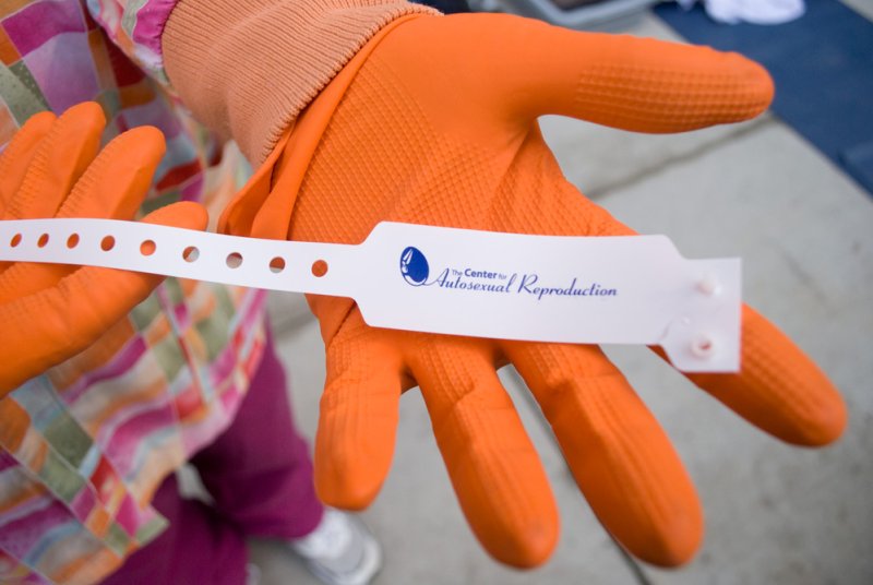 Person in orange gloves and plaid shirt holding a white wristband that reads "Center for Autosexual Reproduction.
