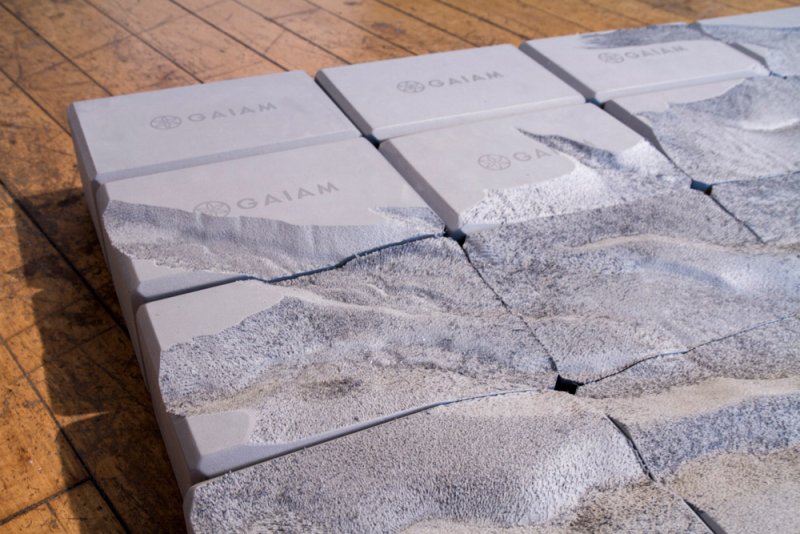 Light gray yoga blocks with "GAIAM" logo, some with textured, eroded surfaces, arranged on a wooden floor.