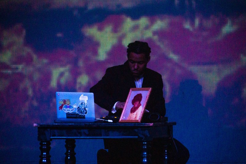 Man in dark suit at a table with a laptop and framed photo, against a blue and purple digital projection.