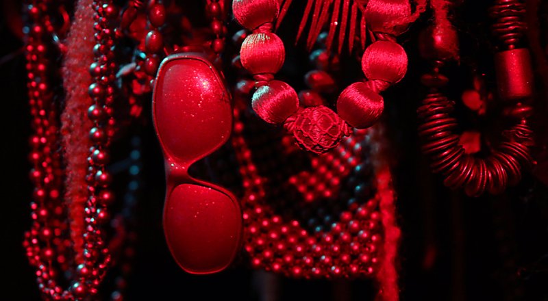 Close-up of red beaded necklaces, sunglasses, and woven textures, illuminated by deep red light against a dark background.