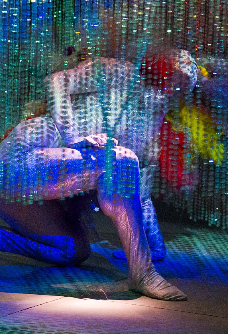 A figure in a shimmering, textured suit kneels, illuminated by blue and purple light with a grid overlay.