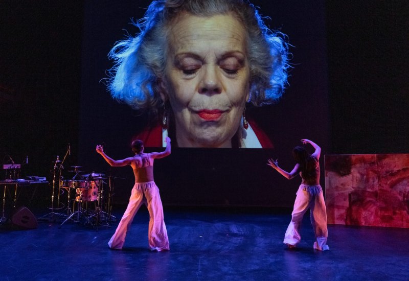 Performance art piece featuring two dancers in loose pink pants, set against a backdrop of a projected close-up of an older woman's face, with musical instruments visible on the side.