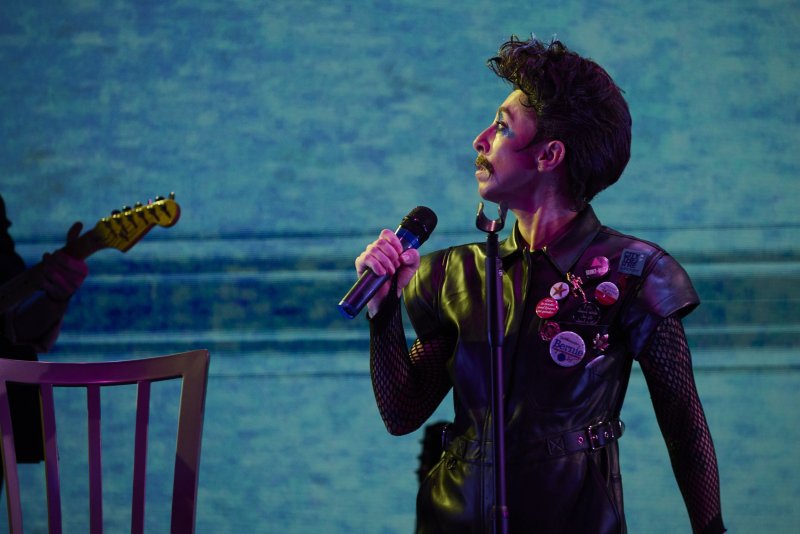 Performer in black leather, fishnet, and makeup sings into a microphone, with a guitarist and blue textured background.