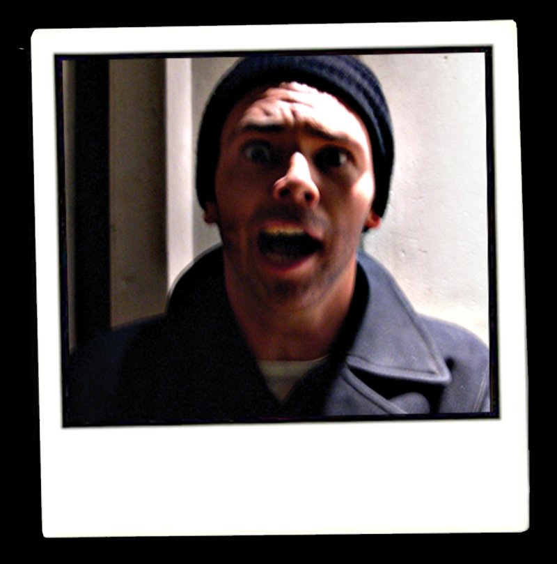 Polaroid-style photo of a man in a black beanie and coat, mouth open in surprise, against a light wall.